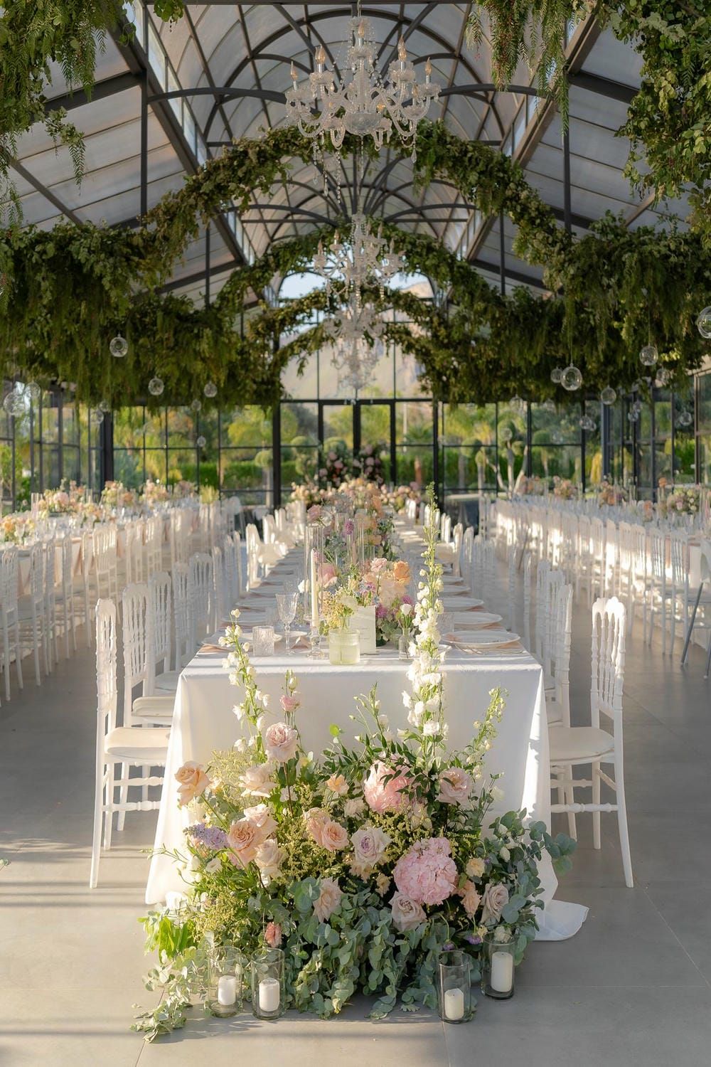 Elegant greenhouse wedding reception setup with crystal chandeliers, white chairs, and lush floral arrangements.