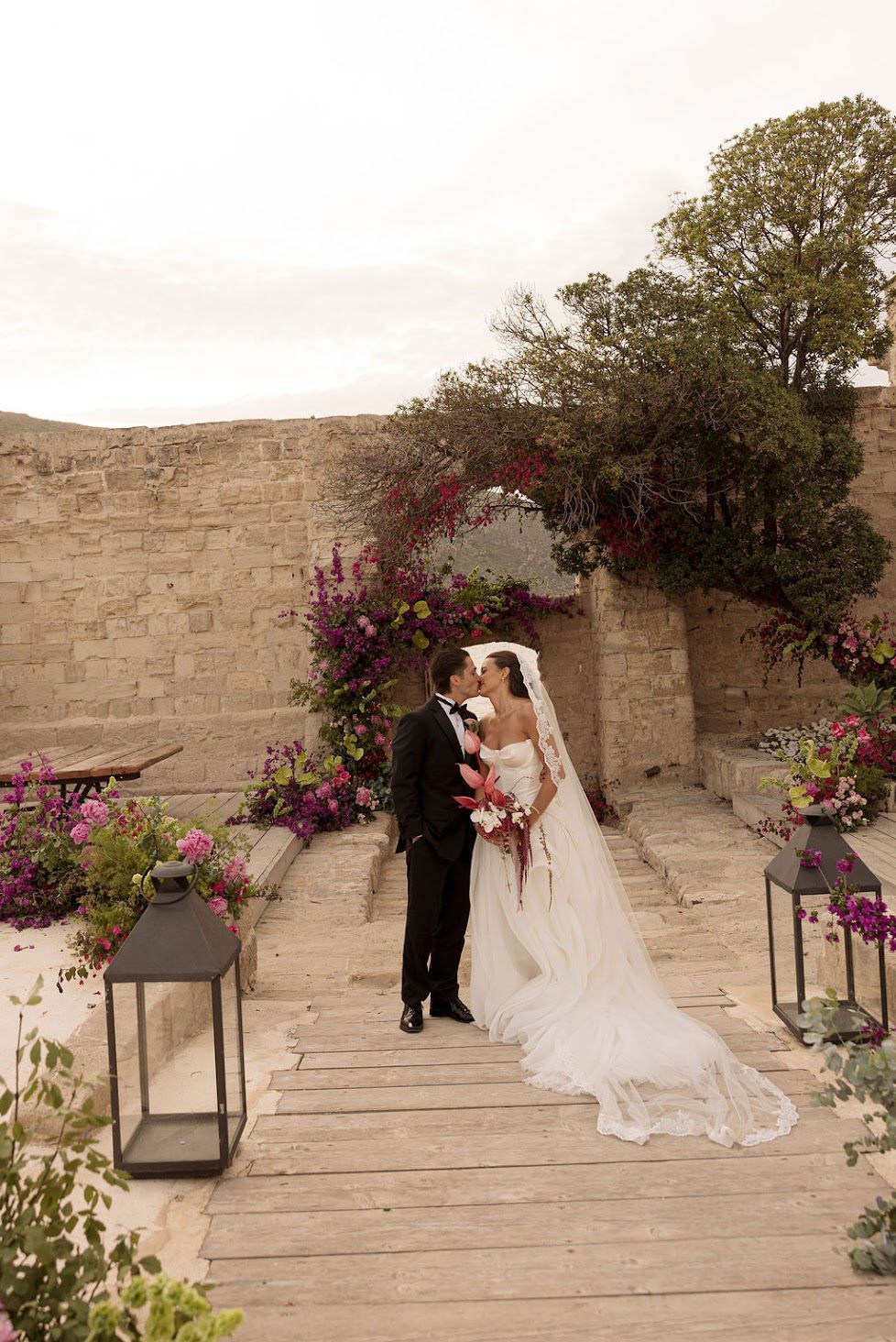 A romantic wedding scene unfolds on stone steps adorned with vibrant purple flowers and lantern decorations.