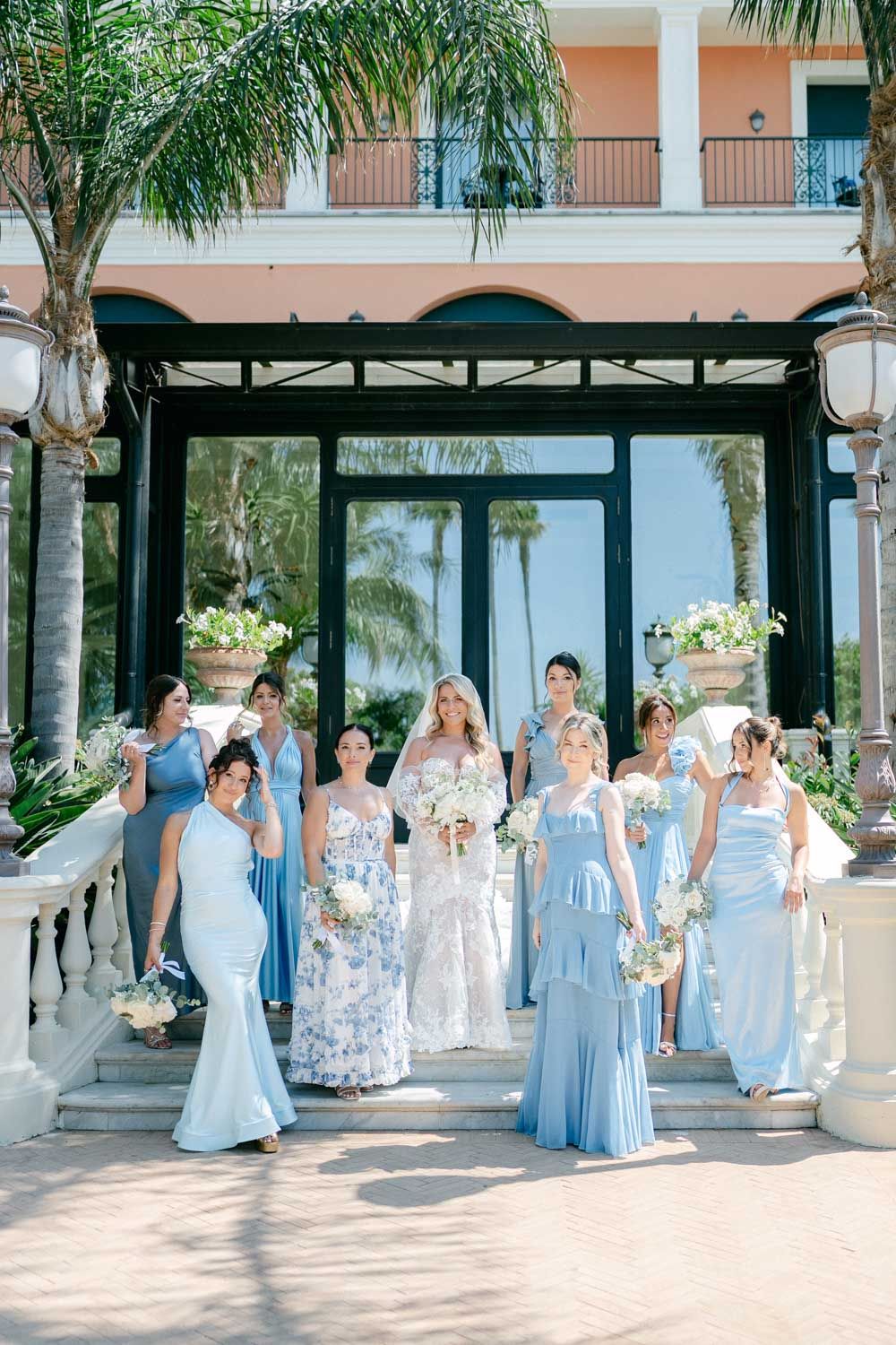 A wedding party in light blue bridesmaid dresses and floral gown pose at a tropical venue with palm trees and columns, Hotel Sorrento
