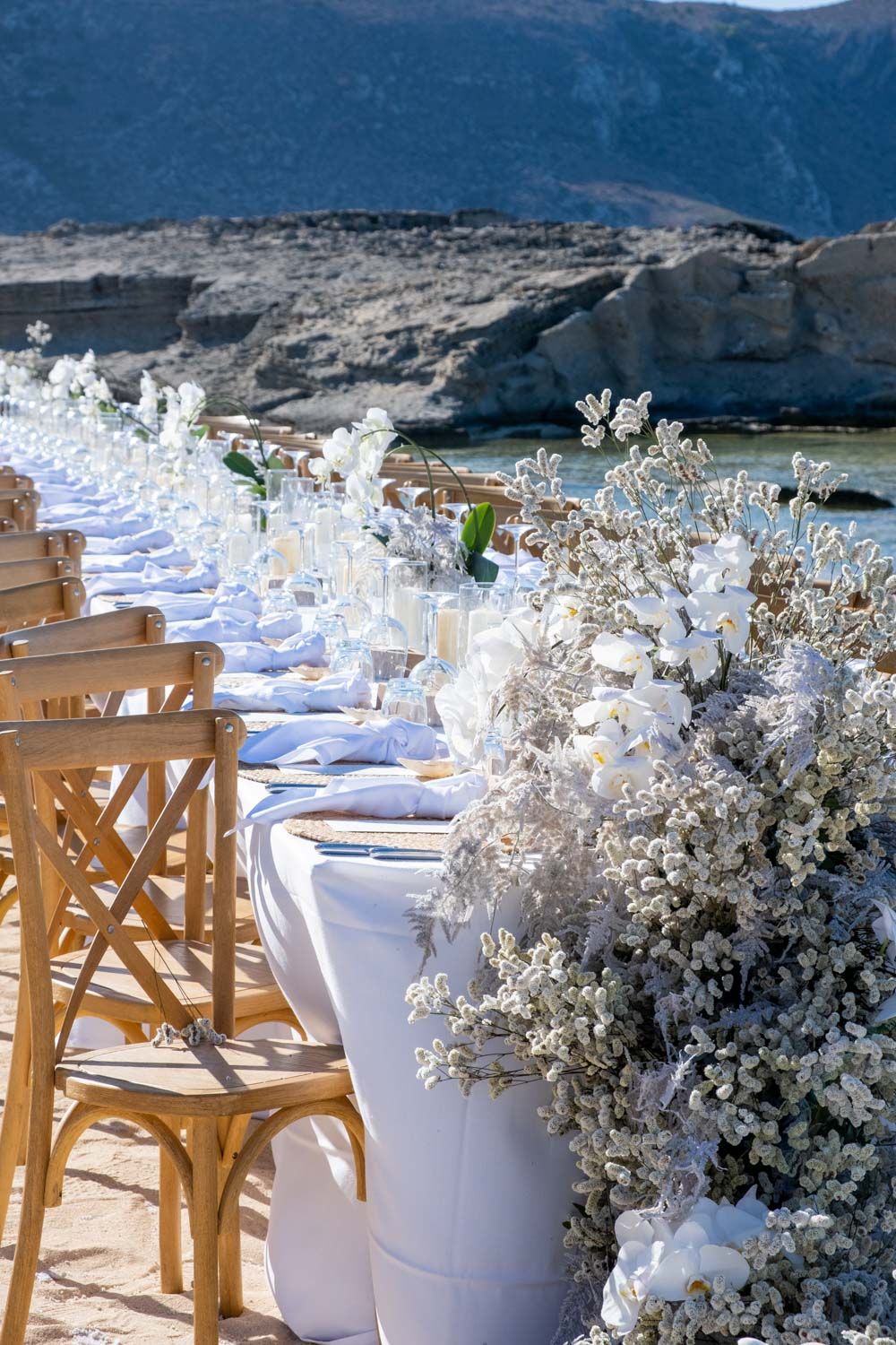 Outdoor wedding reception setup with white linens and wooden chairs against a mountain backdrop.