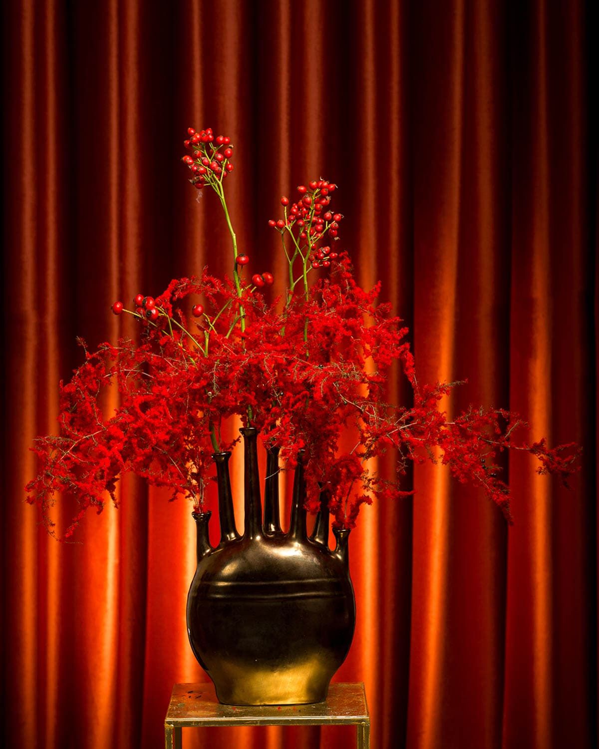 Dramatic red floral arrangement in a black vessel against a rich red curtain background.