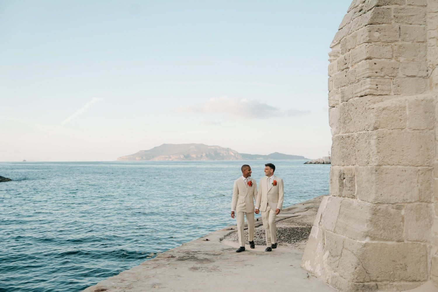 Wedding in Favignana a couple walk on a stone ledge overlooking a serene Mediterranean coastline with turquoise waters.