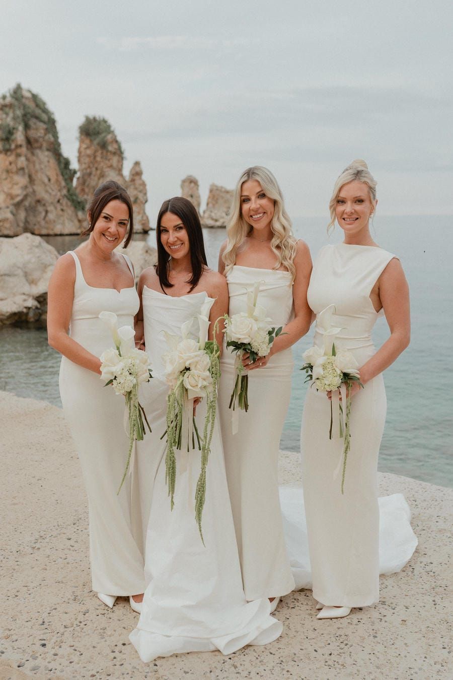 A group of bridesmaids in elegant white dresses hold white floral bouquets while posing together on a beach.