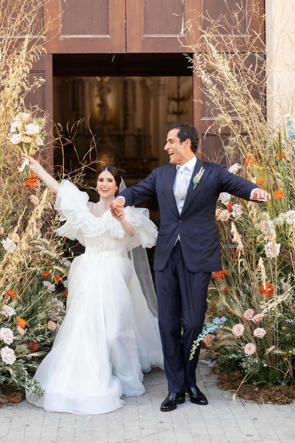 A joyful couple celebrates outside a doorway decorated with autumn flowers and dried grasses.