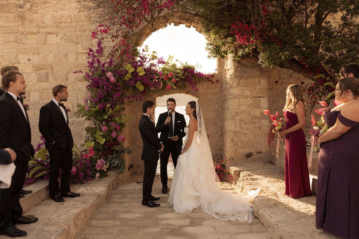 Wedding party gathers in a stone courtyard with vibrant pink bougainvillea flowers creating a romantic atmosphere, Camparia, Favignana.