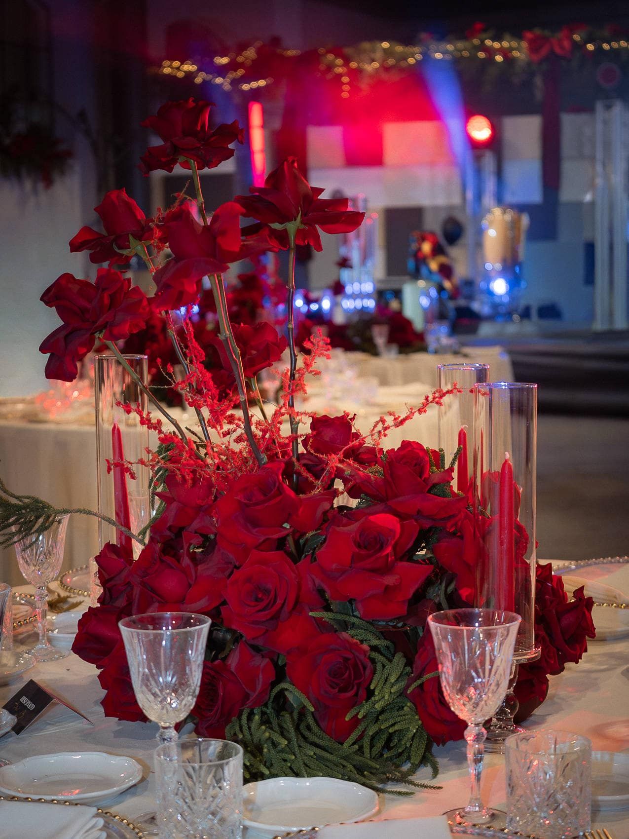 Elegant wedding reception table setting with red rose centerpieces, crystal glassware, and dramatic lighting.