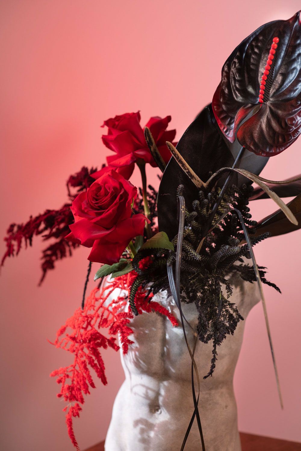 Red roses and dark foliage arranged on a white sculptural bust against a pink background.