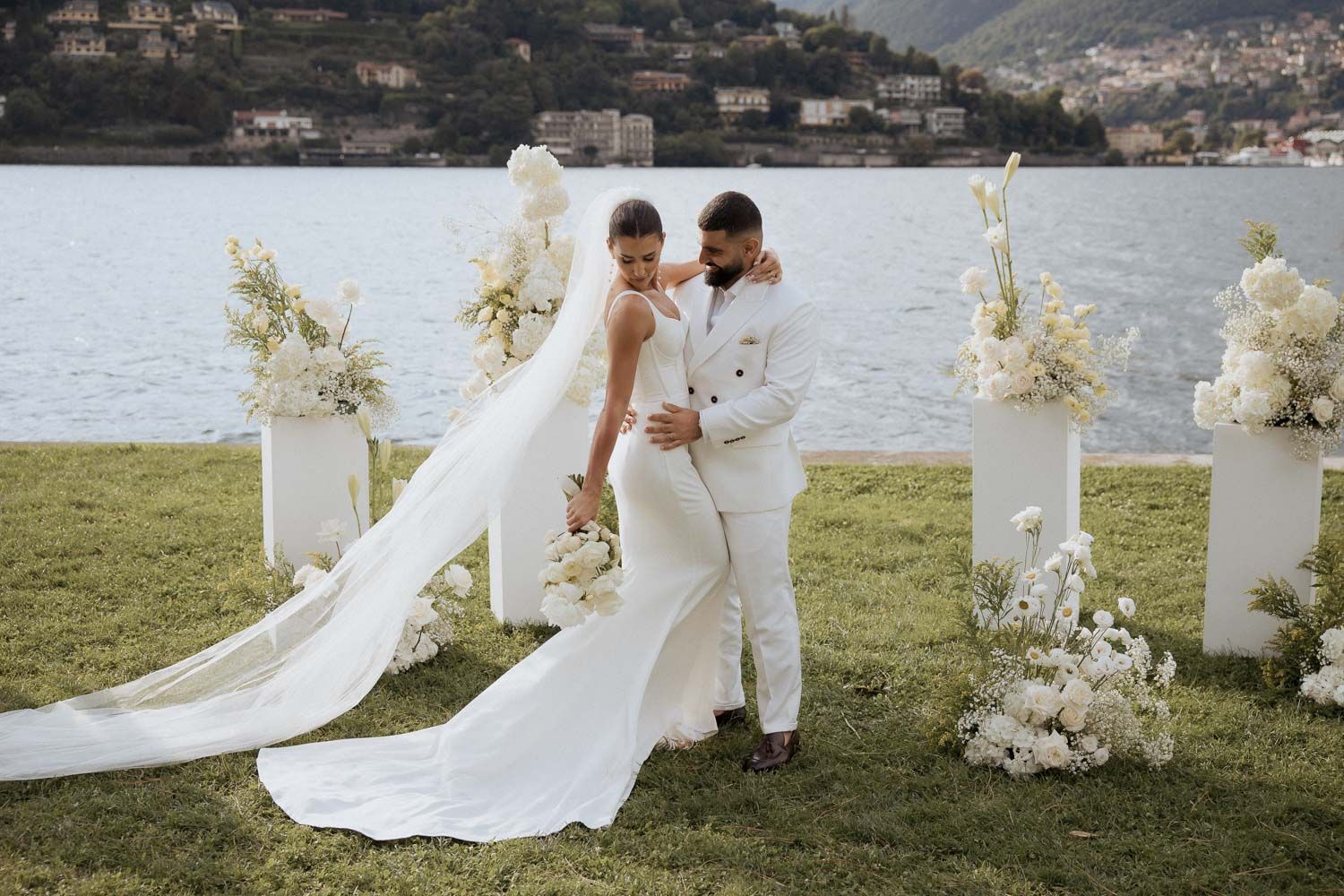 A romantic wedding couple embraces by Lake Como in Italy with mountains and scenic views in the background.