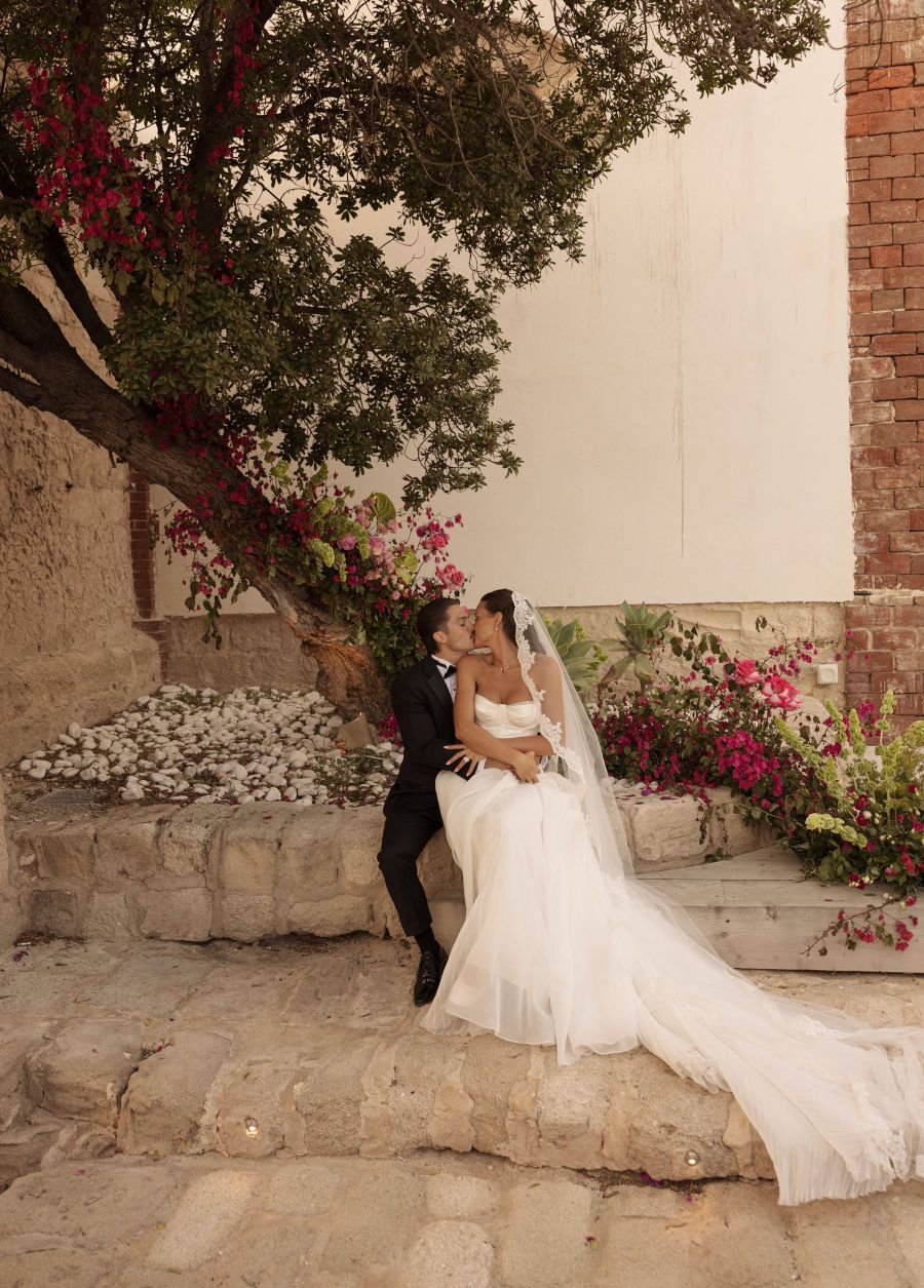 A wedding couple poses against a stone wall with blooming pink bougainvillea flowers in a romantic outdoor setting.