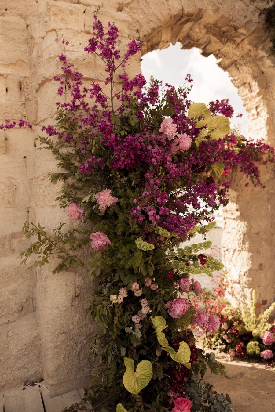 Beautiful pink bougainvillea flowers cascade down a stone wall through an arched window opening in Mediterranean style.