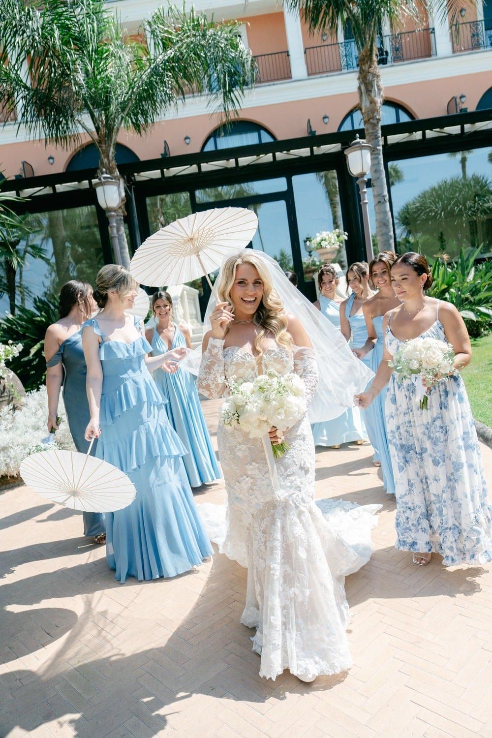 Wedding party members in light blue and floral dresses pose with white parasols on a sunny patio near palm trees.