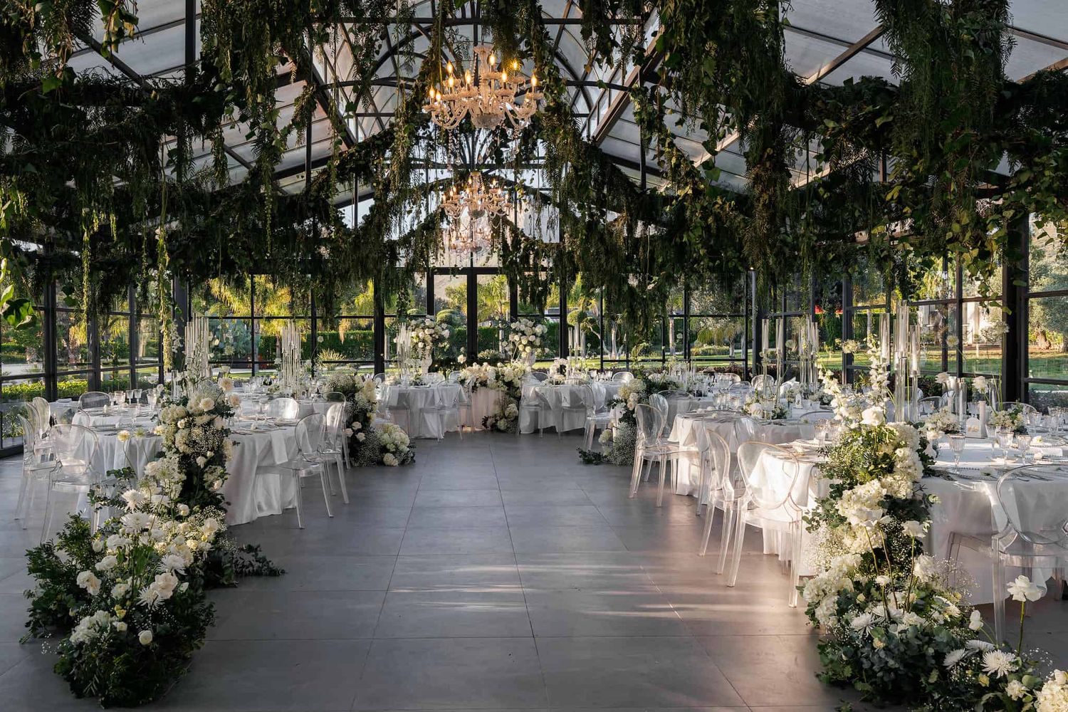 An elegant wedding reception venue with white floral arrangements and crystal chandeliers in a glass-enclosed space.