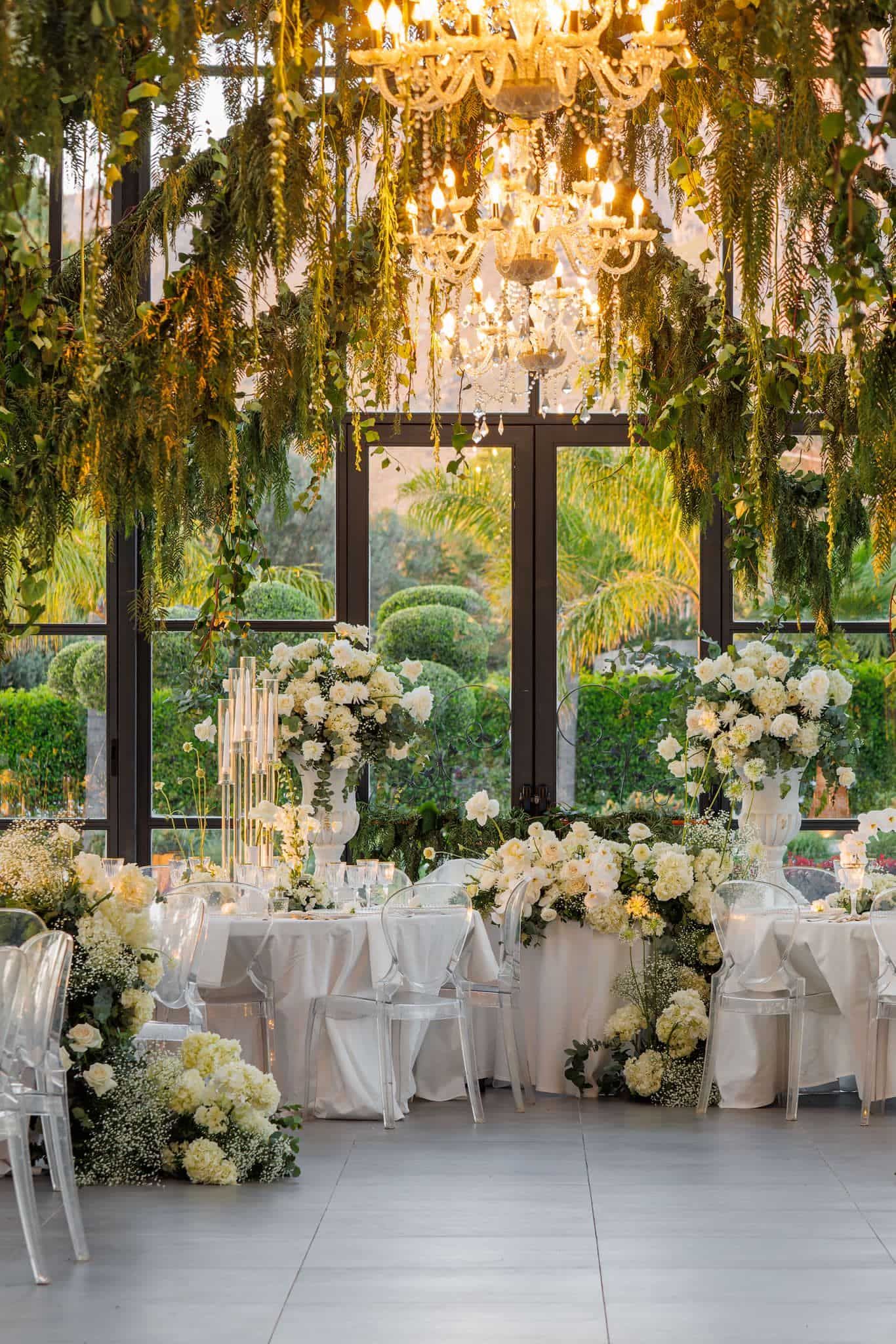 Elegant wedding reception venue with crystal chandeliers, white floral arrangements and round tables in a glass-enclosed space.