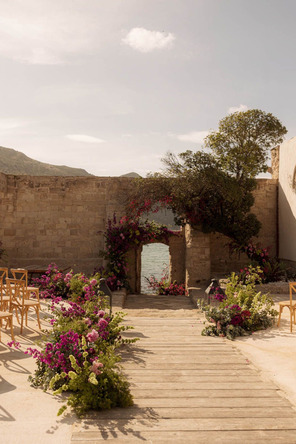 Panoramic view of a desert courtyard with stone walls, wooden steps, and vibrant pink bougainvillea flowers, Camparia, Favignana.
