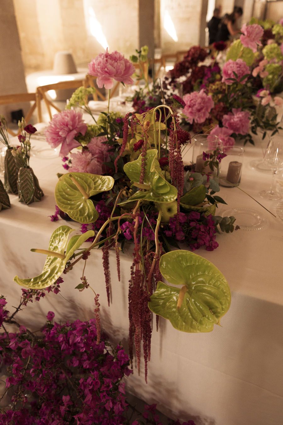 Elegant floral arrangement with pink carnations and green anthurium leaves on a white tablecloth in warm lighting.