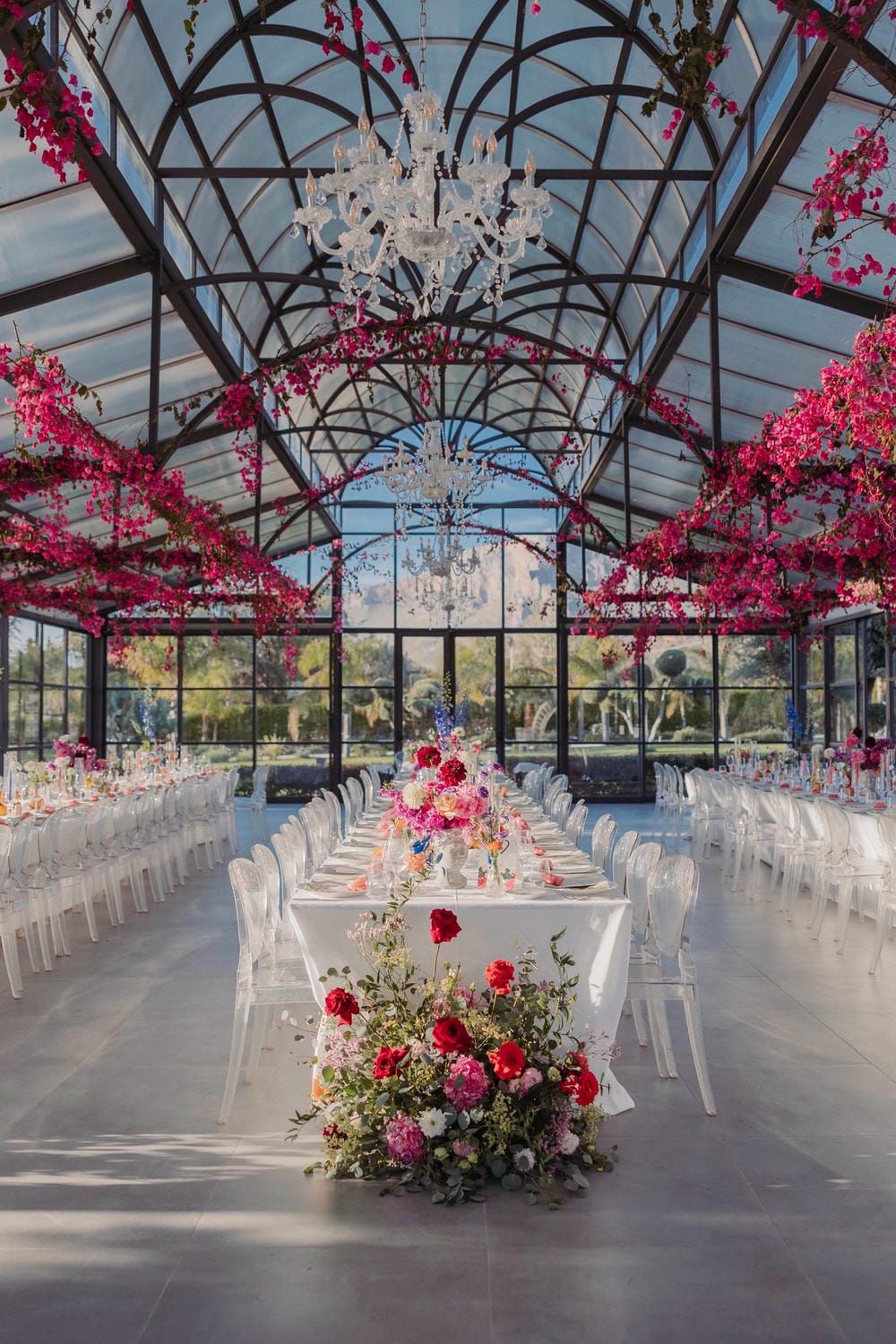 Modern glass greenhouse wedding venue with pink floral decor, crystal chandeliers, and white chairs arranged in rows.