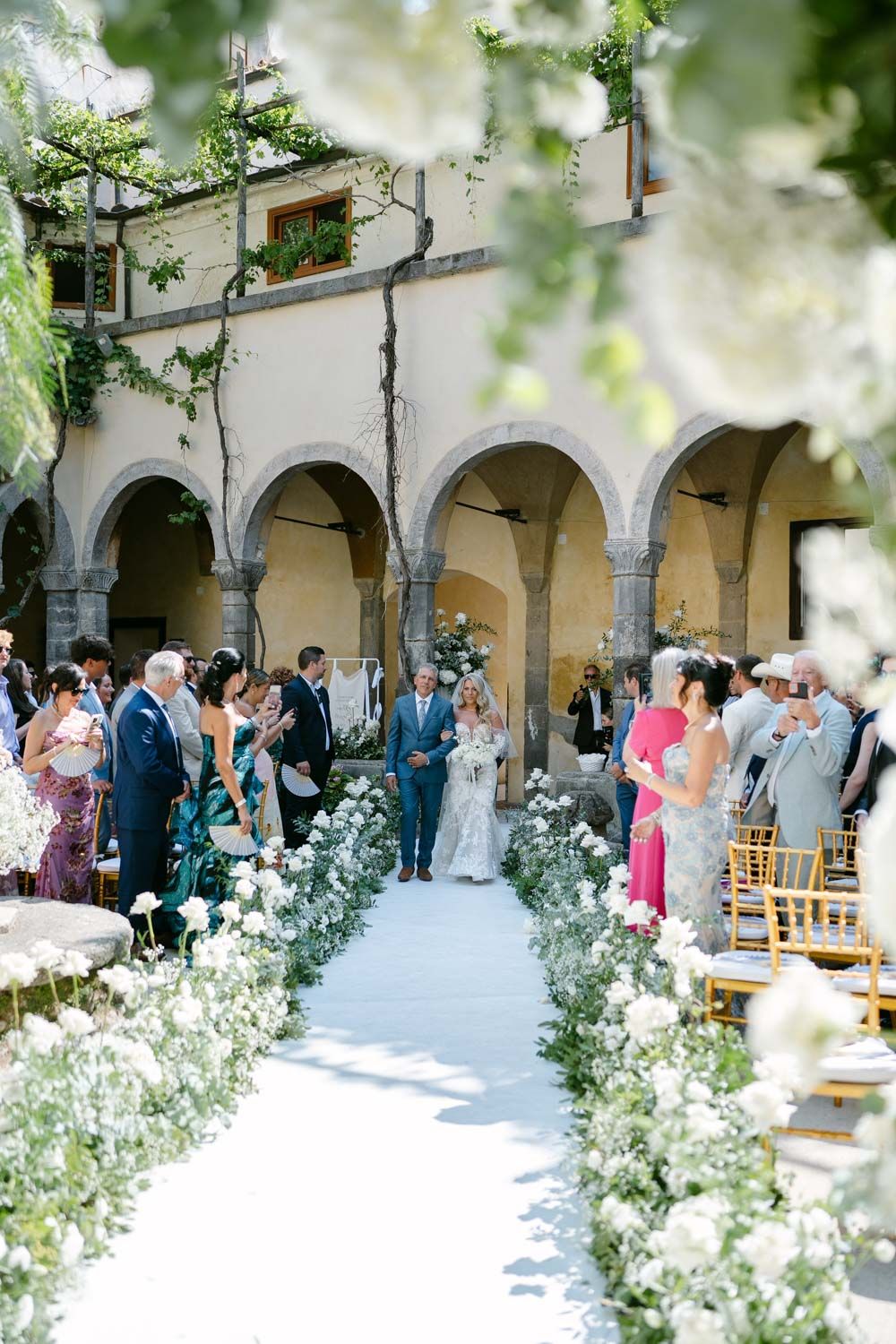 Outdoor wedding ceremony in elegant courtyard with white floral arrangements and arched architecture.