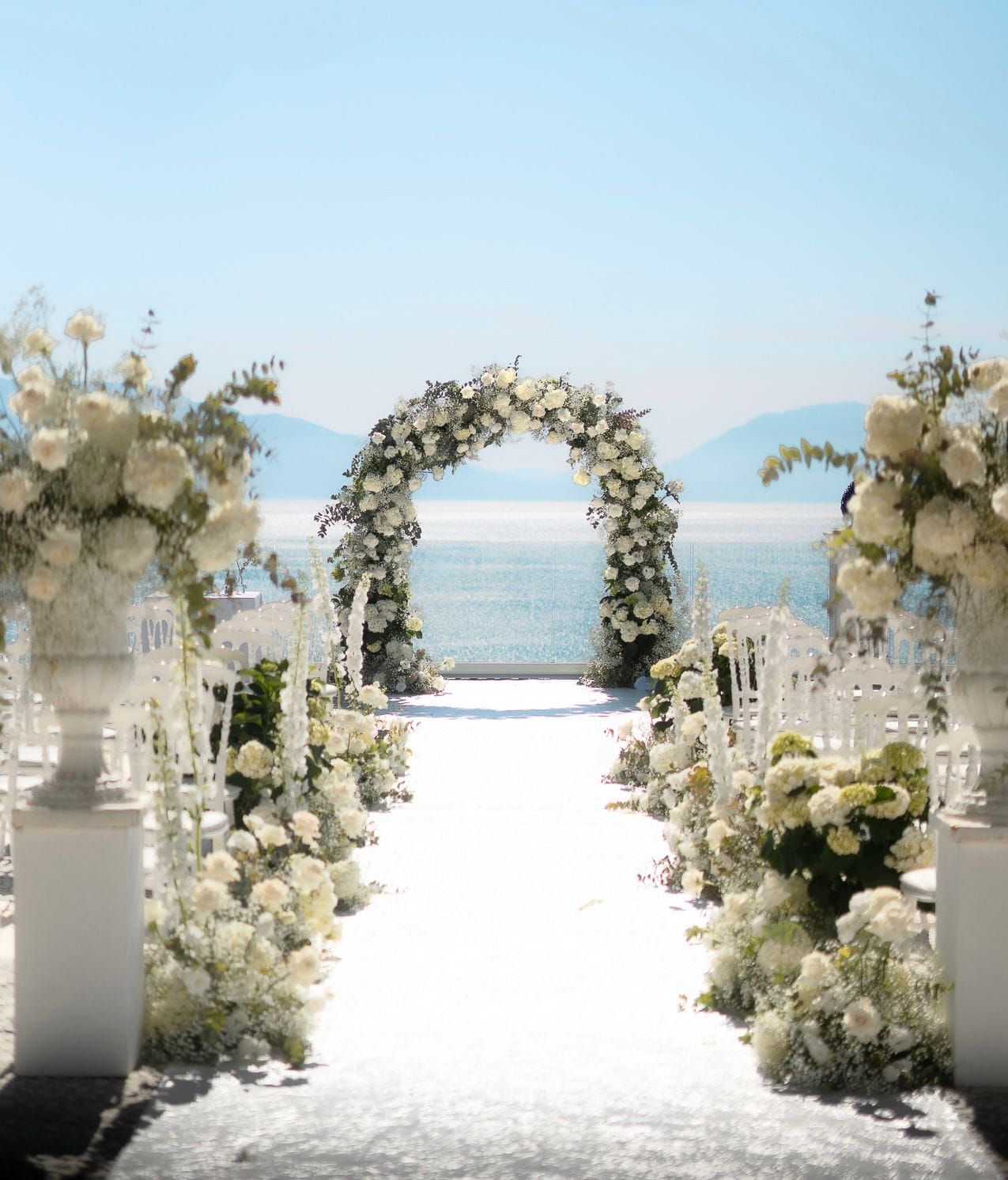 Elegant white floral wedding arch and aisle decorations overlooking an ocean view with white flowers and greenery.