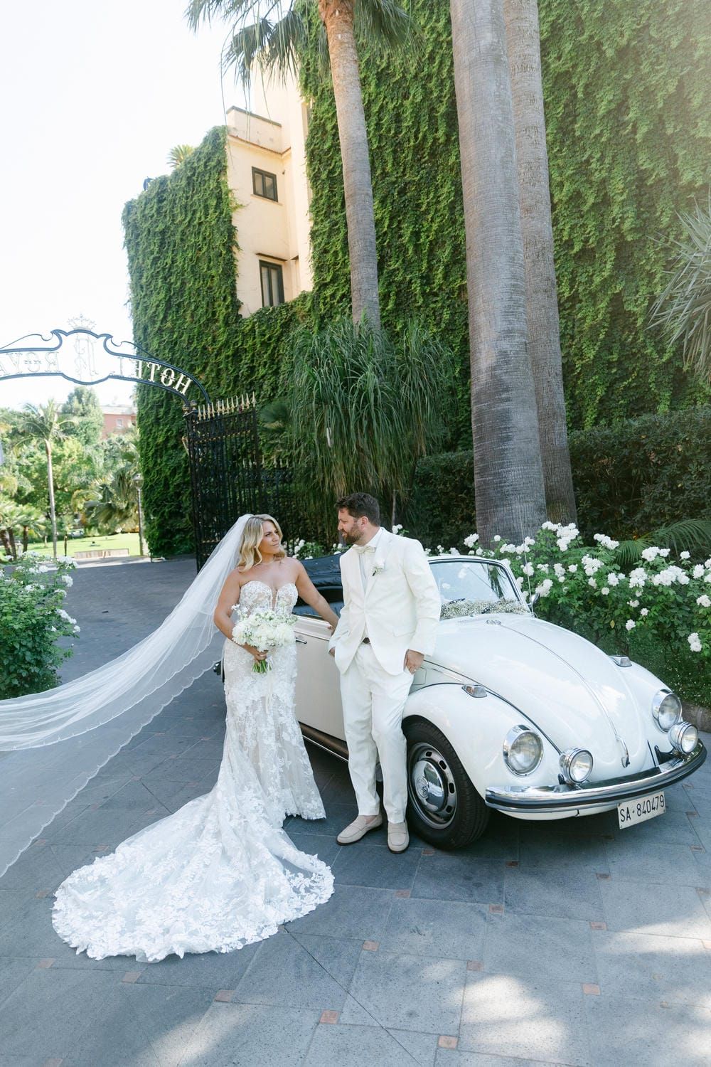 A newlywed couple poses with a classic white Volkswagen Beetle at a lush tropical venue with ivy-covered walls.