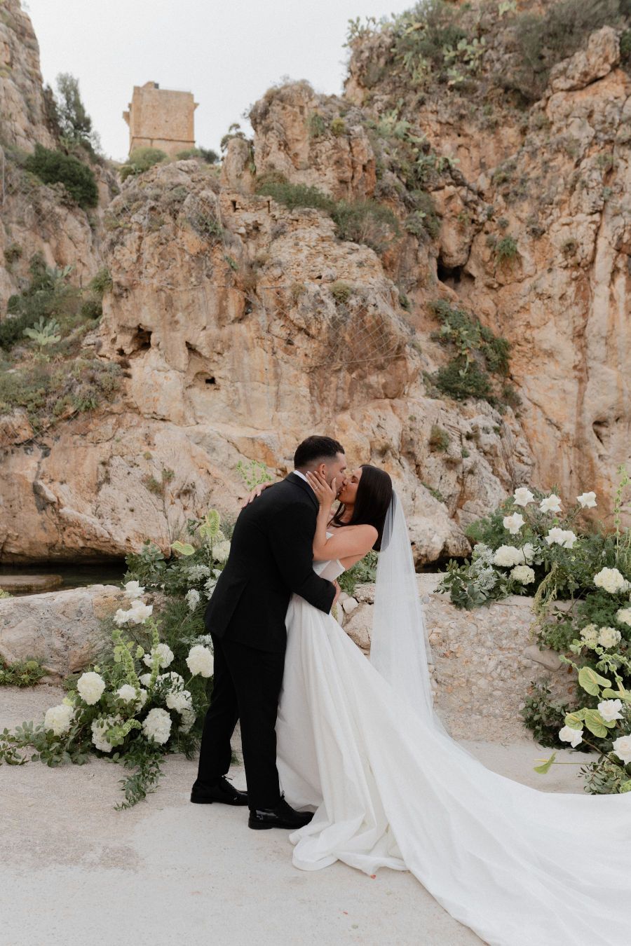 A romantic wedding ceremony set against dramatic rocky cliffs with white floral arrangements and a flowing bridal veil, Scopello, Sicily.