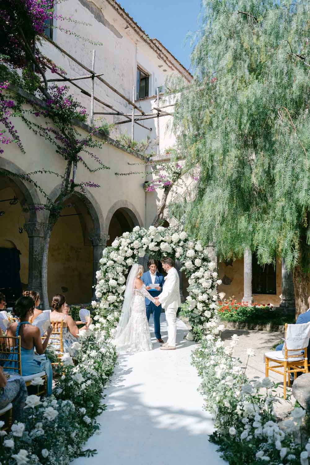 Sun-drenched courtyard wedding ceremony with white floral arch and willow trees.