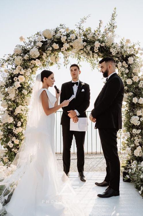 A wedding ceremony takes place under an elegant white floral arch with a scenic ocean backdrop at tenuta cala muletti.