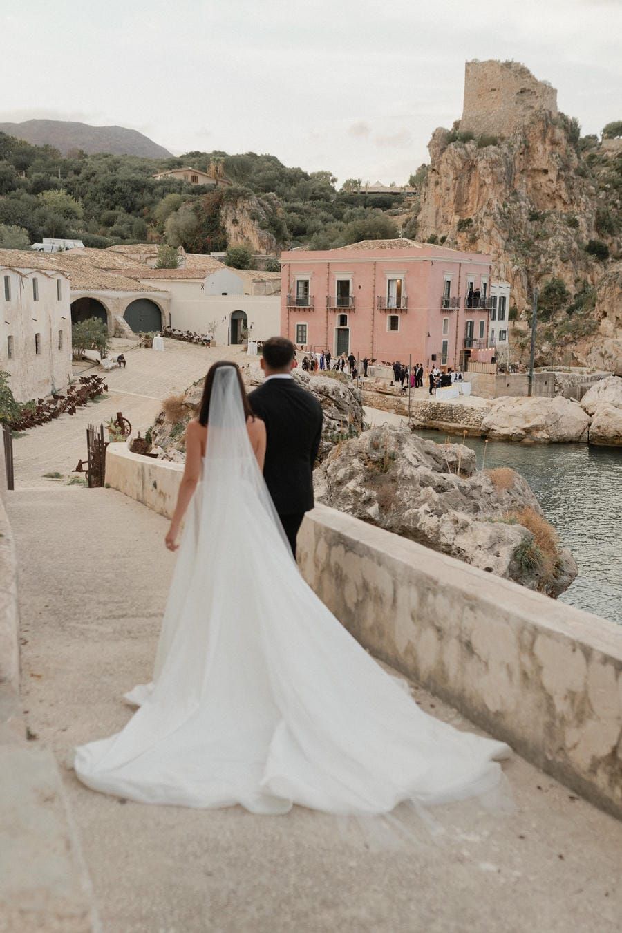 A couple in wedding attire walks along a scenic coastal path with stone walls and Mediterranean architecture.