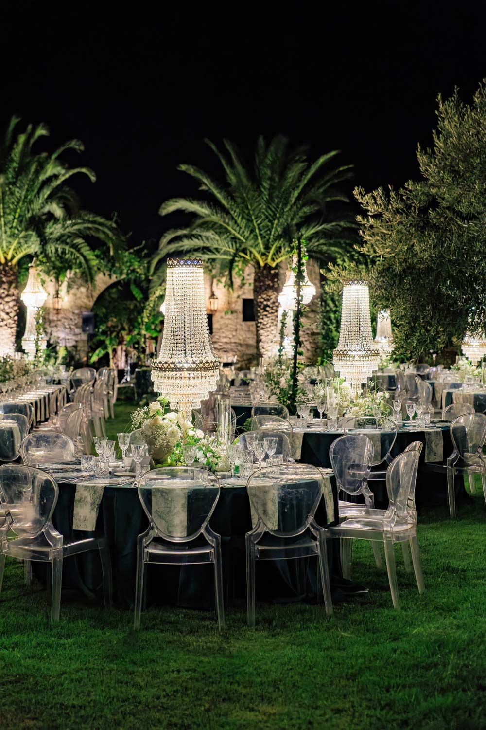 Elegant outdoor wedding reception setup with crystal chandeliers, ghost chairs and palm trees lit up at night.