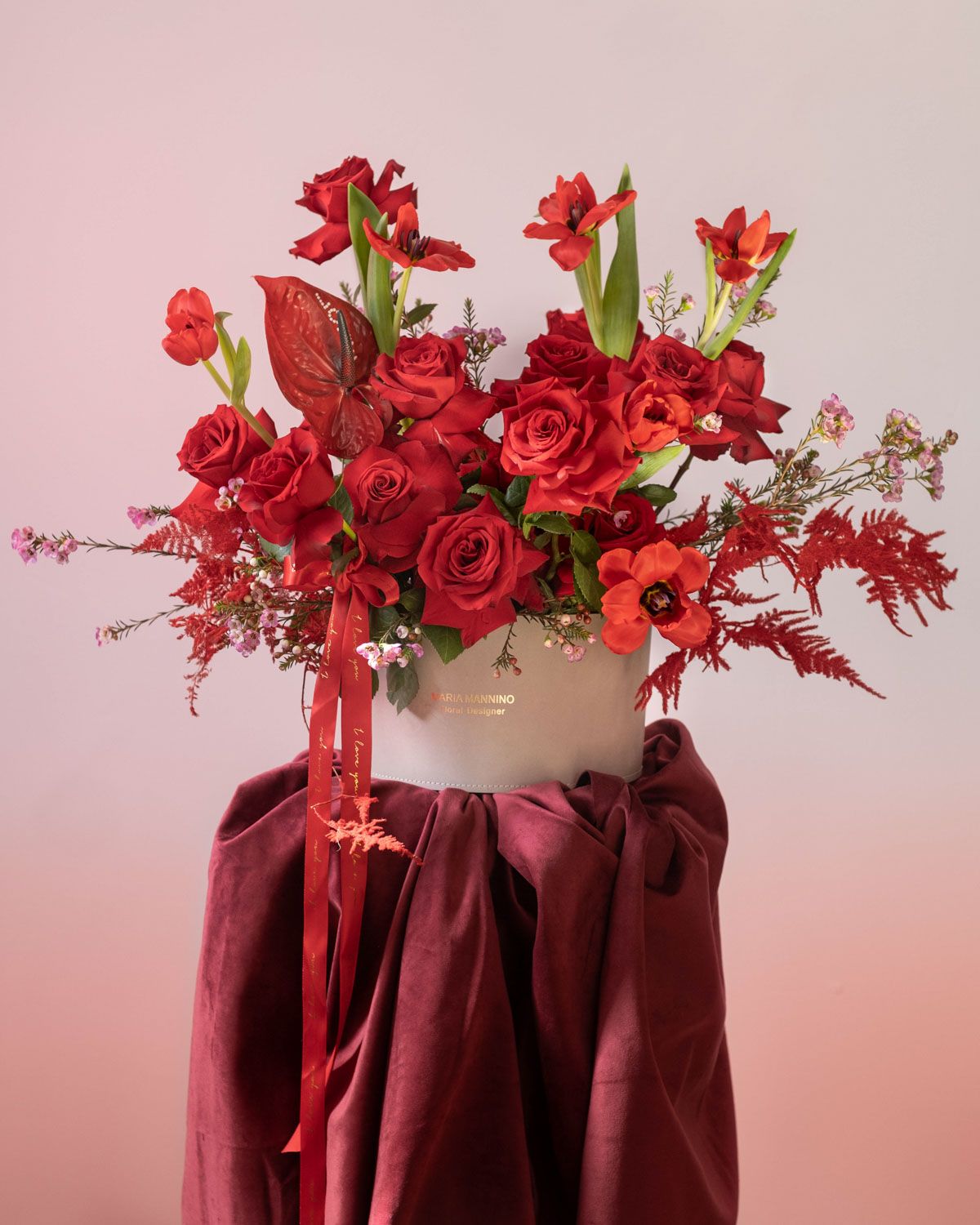 Red roses and flowers arranged on a burgundy draped pedestal with soft pink lighting.