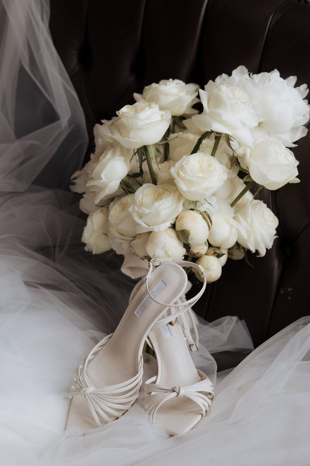 White wedding shoes and elegant white rose bouquet arranged with tulle fabric against dark background.