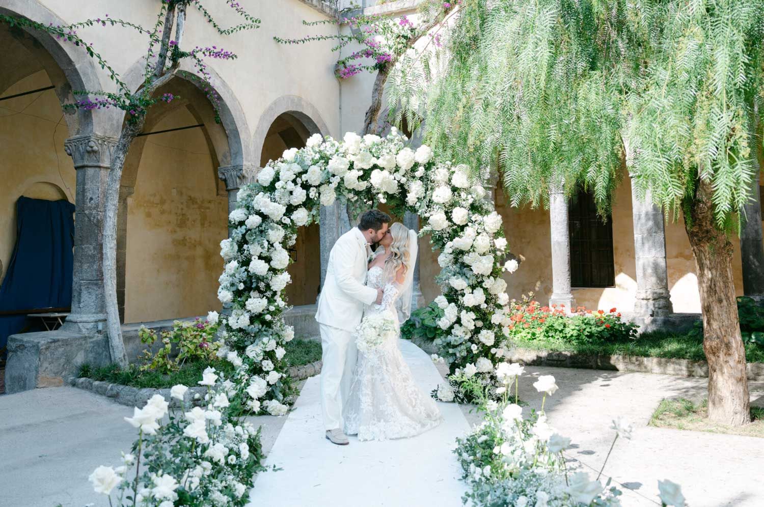 Elegant wedding ceremony setup in a Mediterranean courtyard with white floral arrangements and stone archways.
