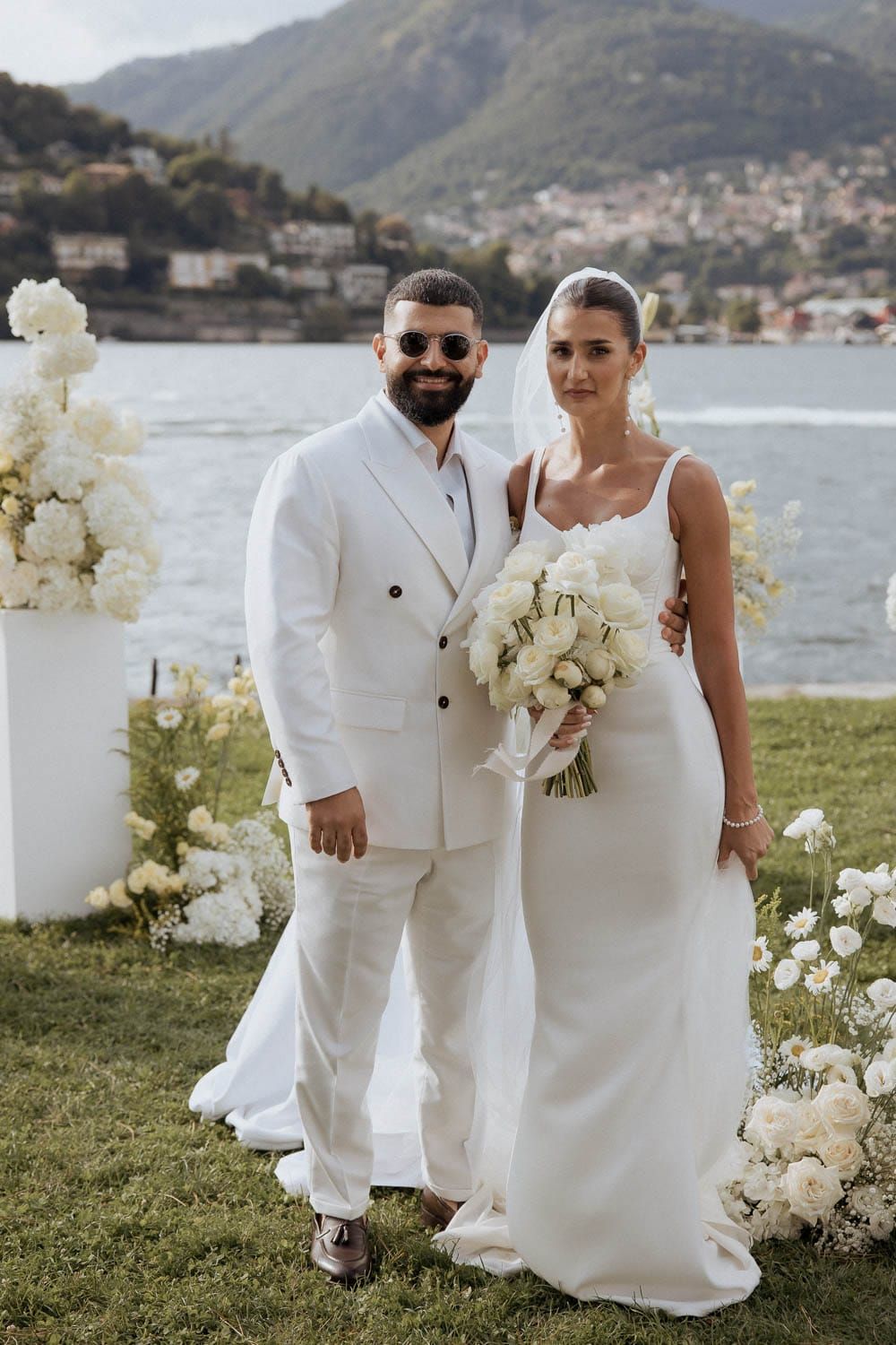 A couple in matching white outfits poses for wedding photos by a scenic lakeside mountain view.