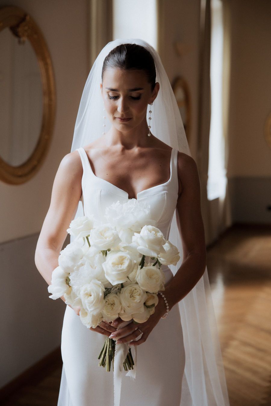 A wedding bouquet of white roses is held against a white bridal gown in an elegant indoor setting.