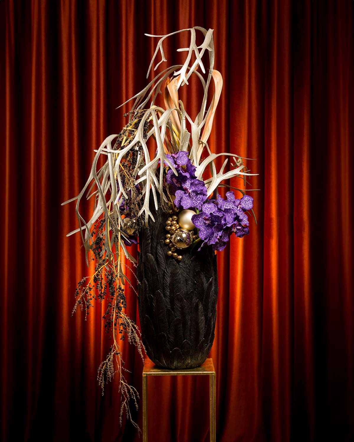 Elegant purple flowers and dried grass arranged in black vases against a dramatic red velvet curtain backdrop.