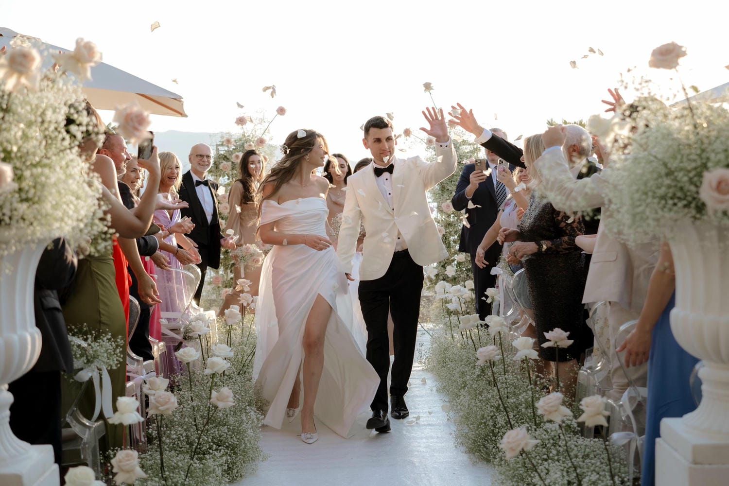 Wedding guests gather around a floral-decorated outdoor ceremony space with white decor and natural greenery.