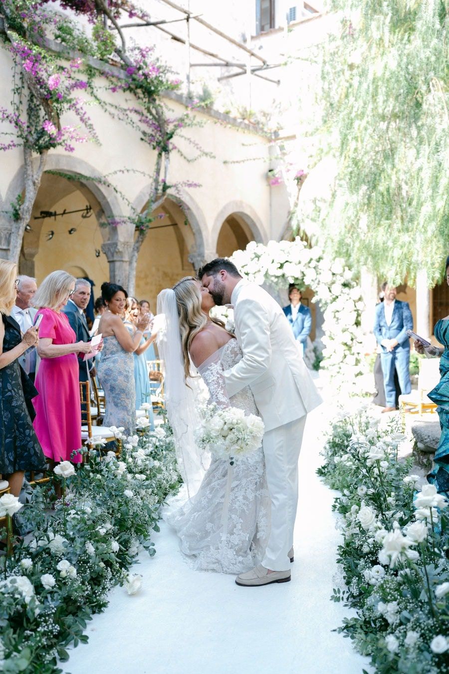Wedding guests dancing together in an elegant courtyard with willow trees and purple flowers under arched walkways.