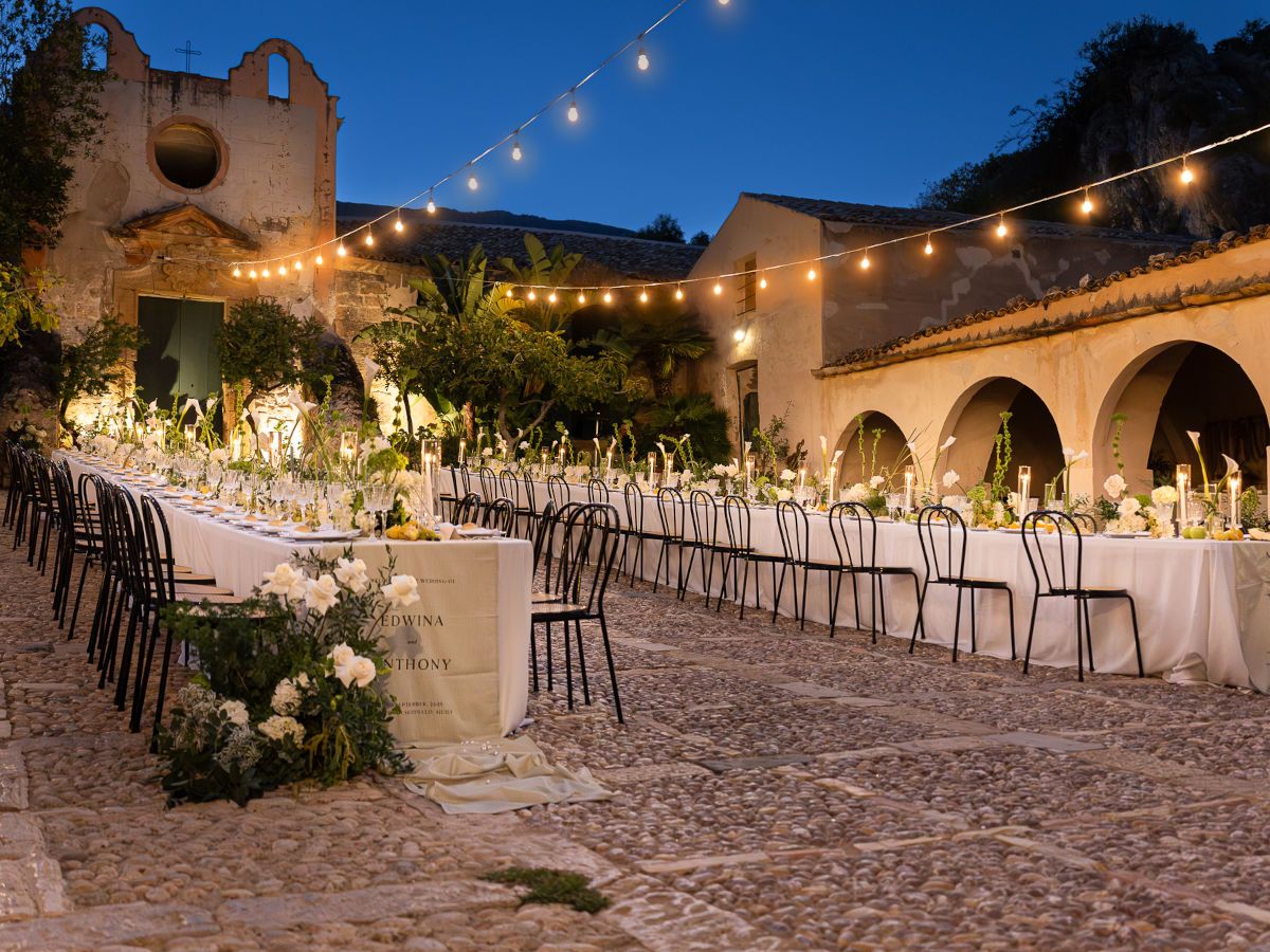 String lights illuminate an outdoor wedding reception venue with long tables and chairs on a stone patio at dusk.