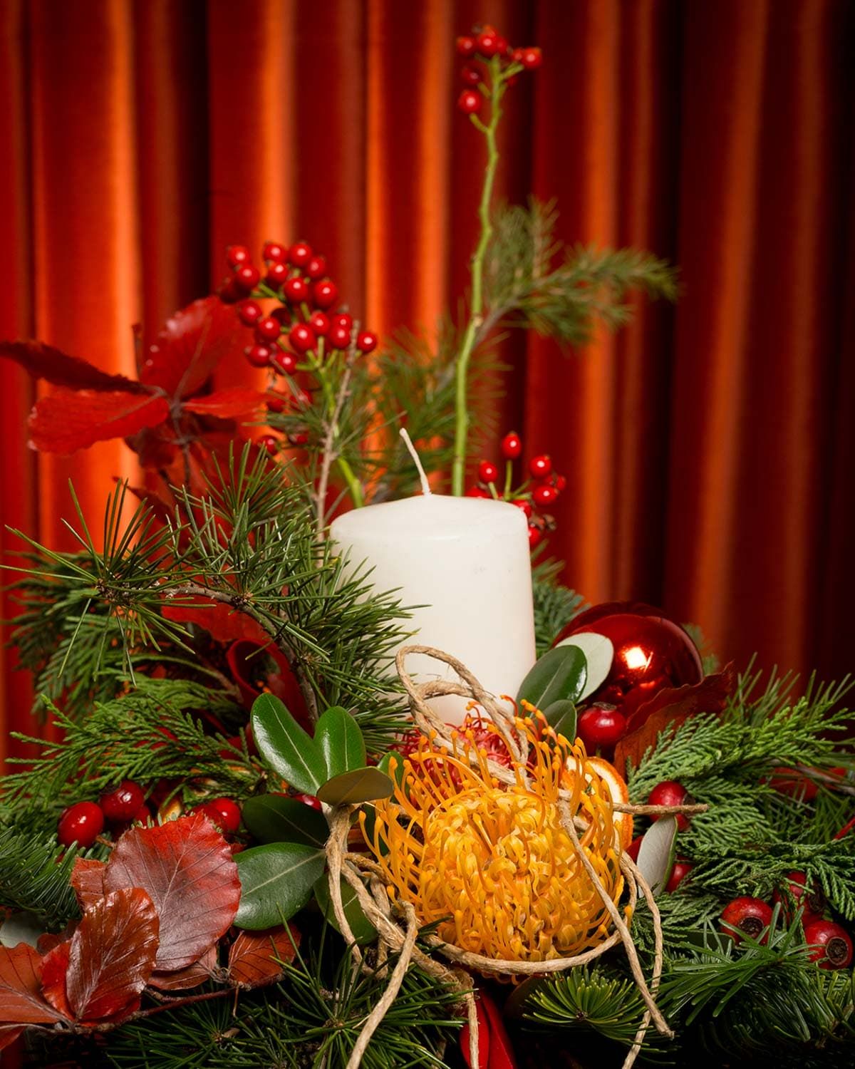 Holiday centerpiece with white candle, pine branches, and red berries against red drapery.