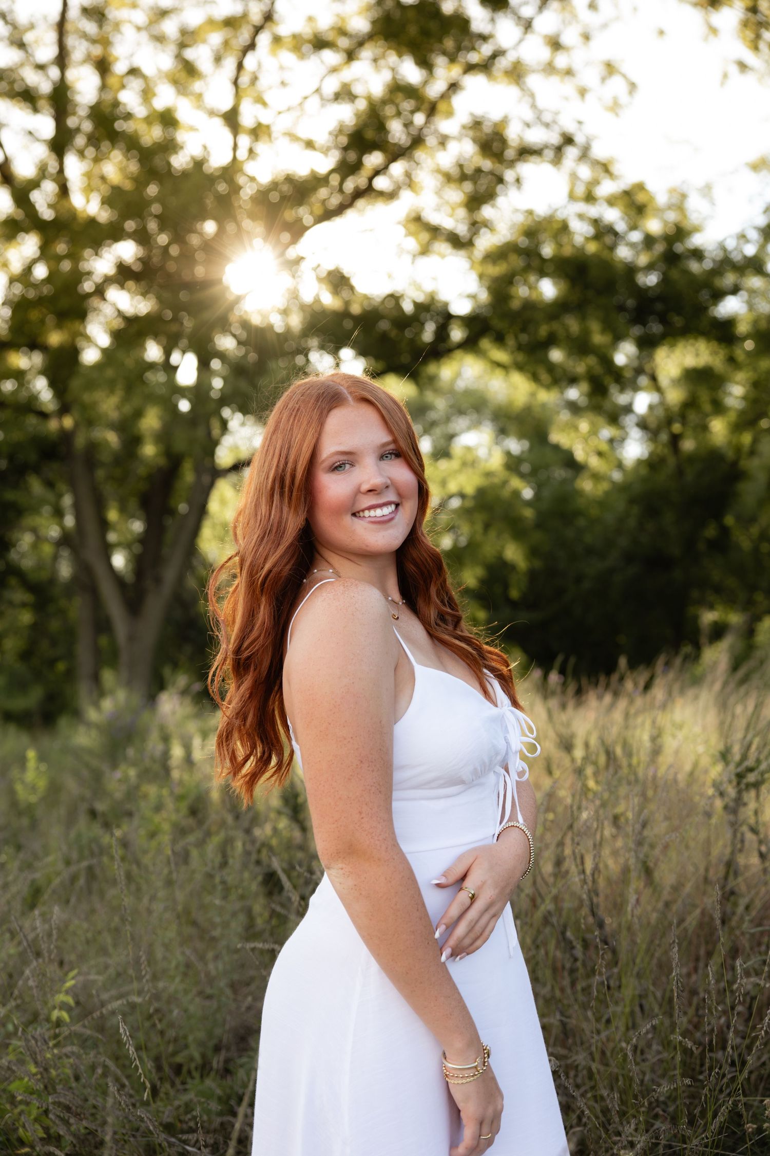 A woman in white dress smiles radiantly during a sunset photoshoot in a natural meadow setting.
