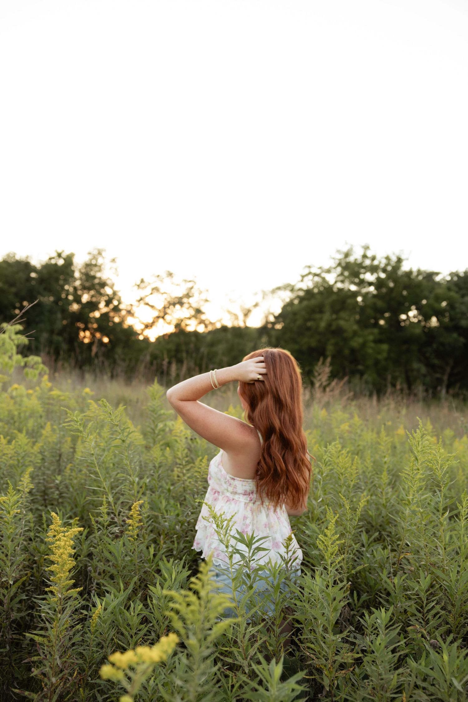 A woman in white dress stands among tall yellow wildflowers at sunset.
