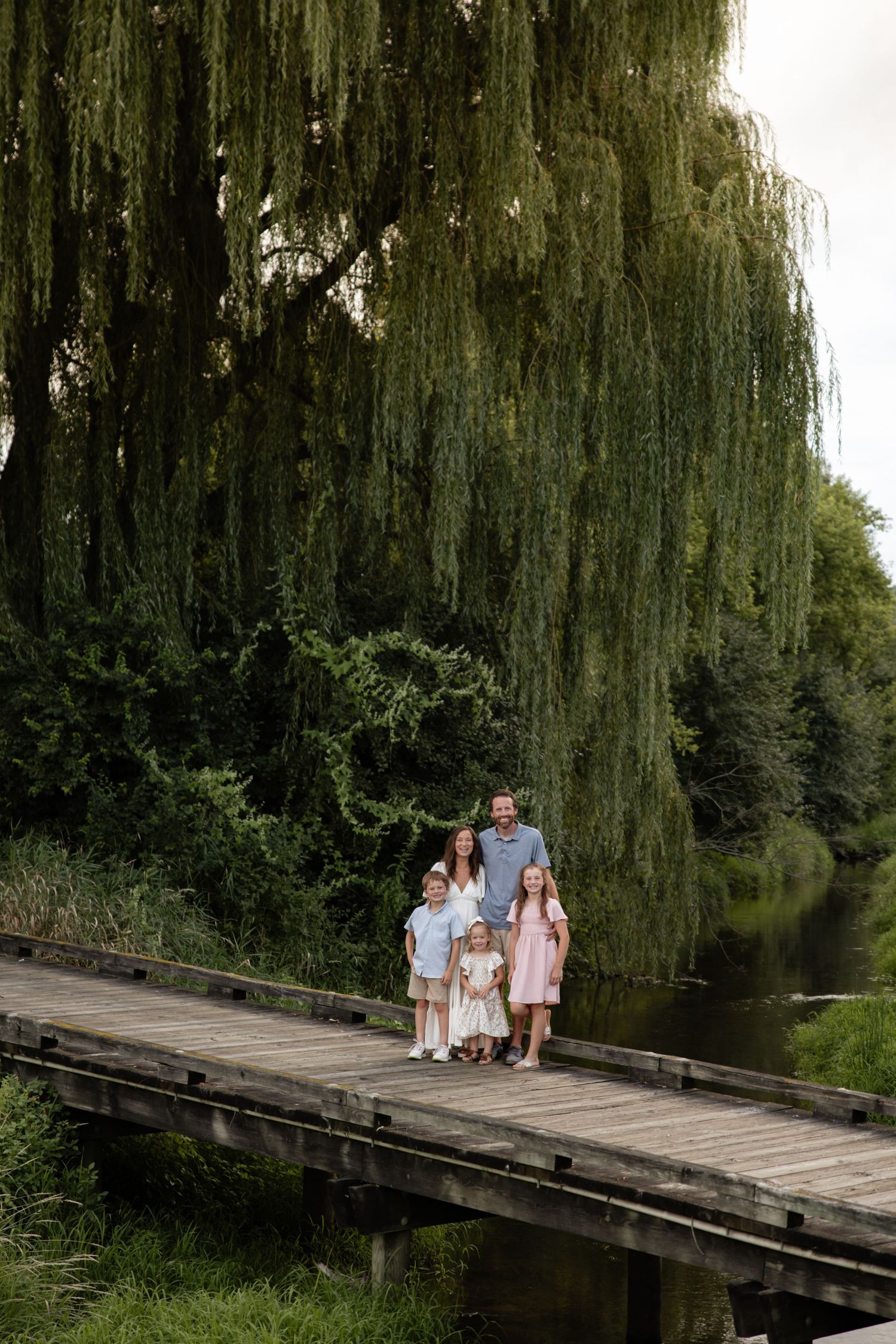 Family walking together on a wooden bridge under a dramatic weeping willow tree.