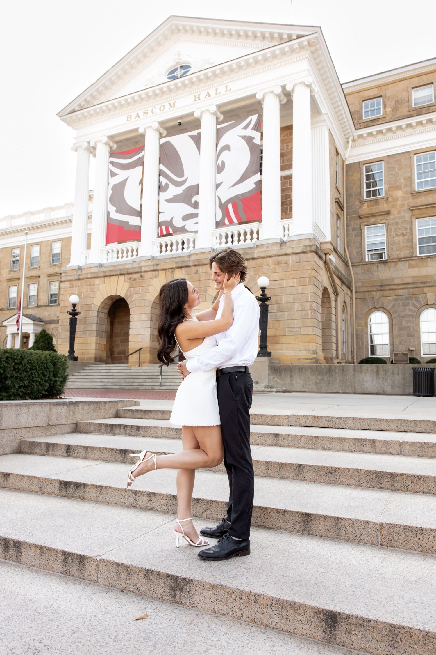 A couple shares a romantic moment on the steps of a historic university building with white columns and red banners.