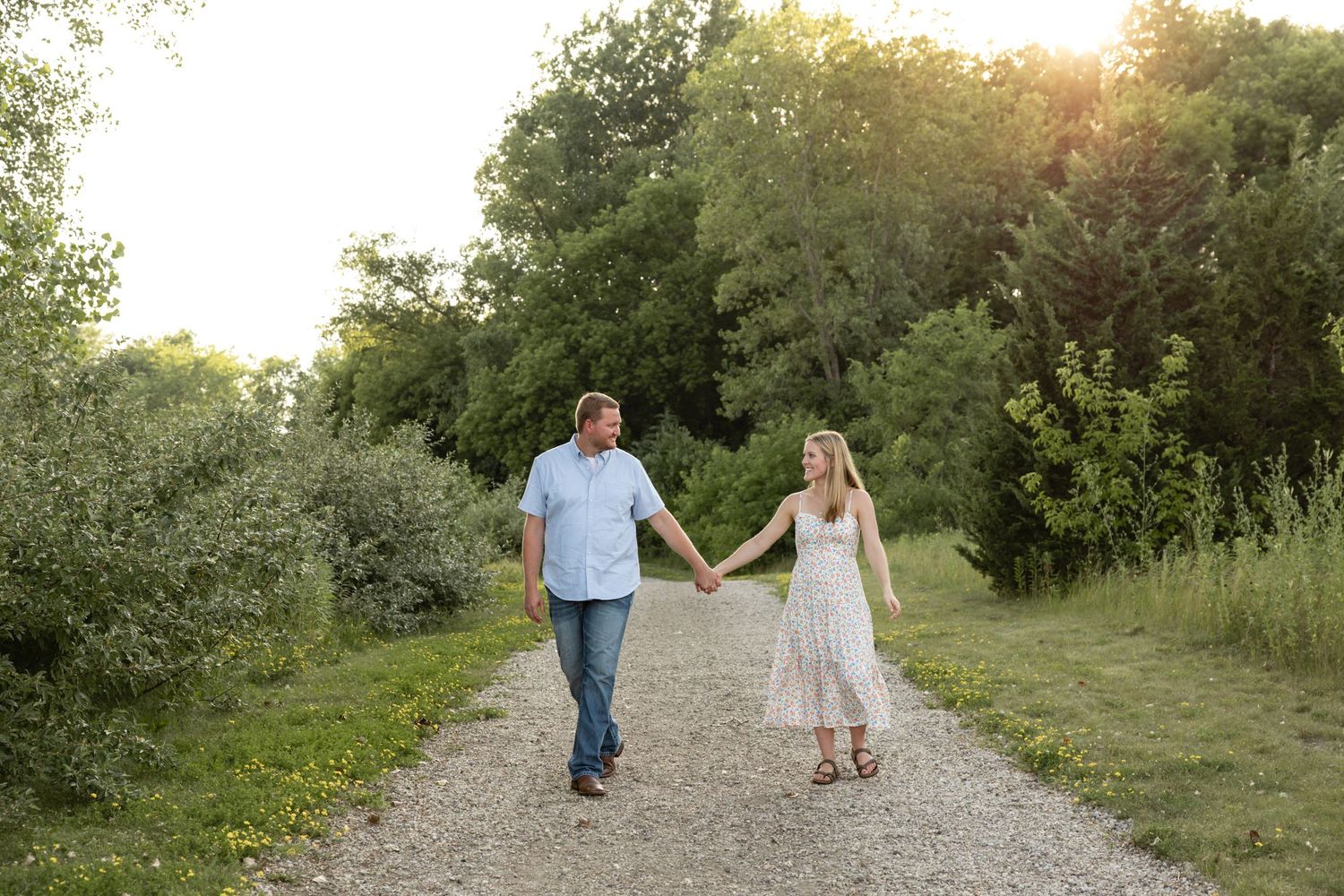 A couple walks hand in hand down a nature path during golden hour surrounded by lush greenery.