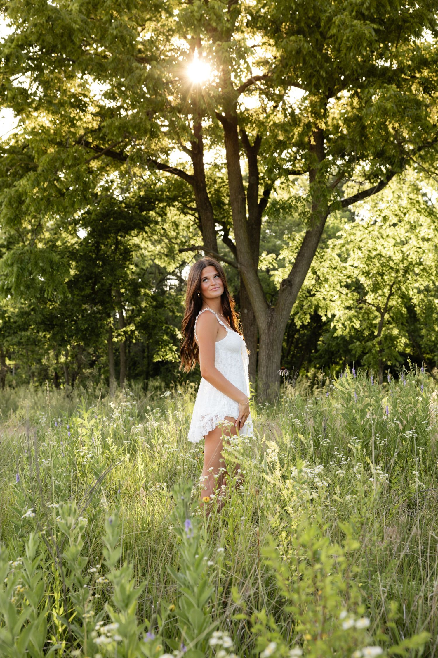 Woman in white dress poses gracefully in a summer meadow with tall grass and trees.