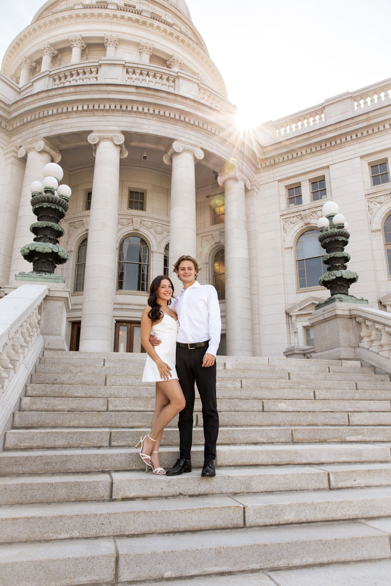 A couple stands on the grand steps of the Wisconsin State Capitol with sunlight peeking through the dome.