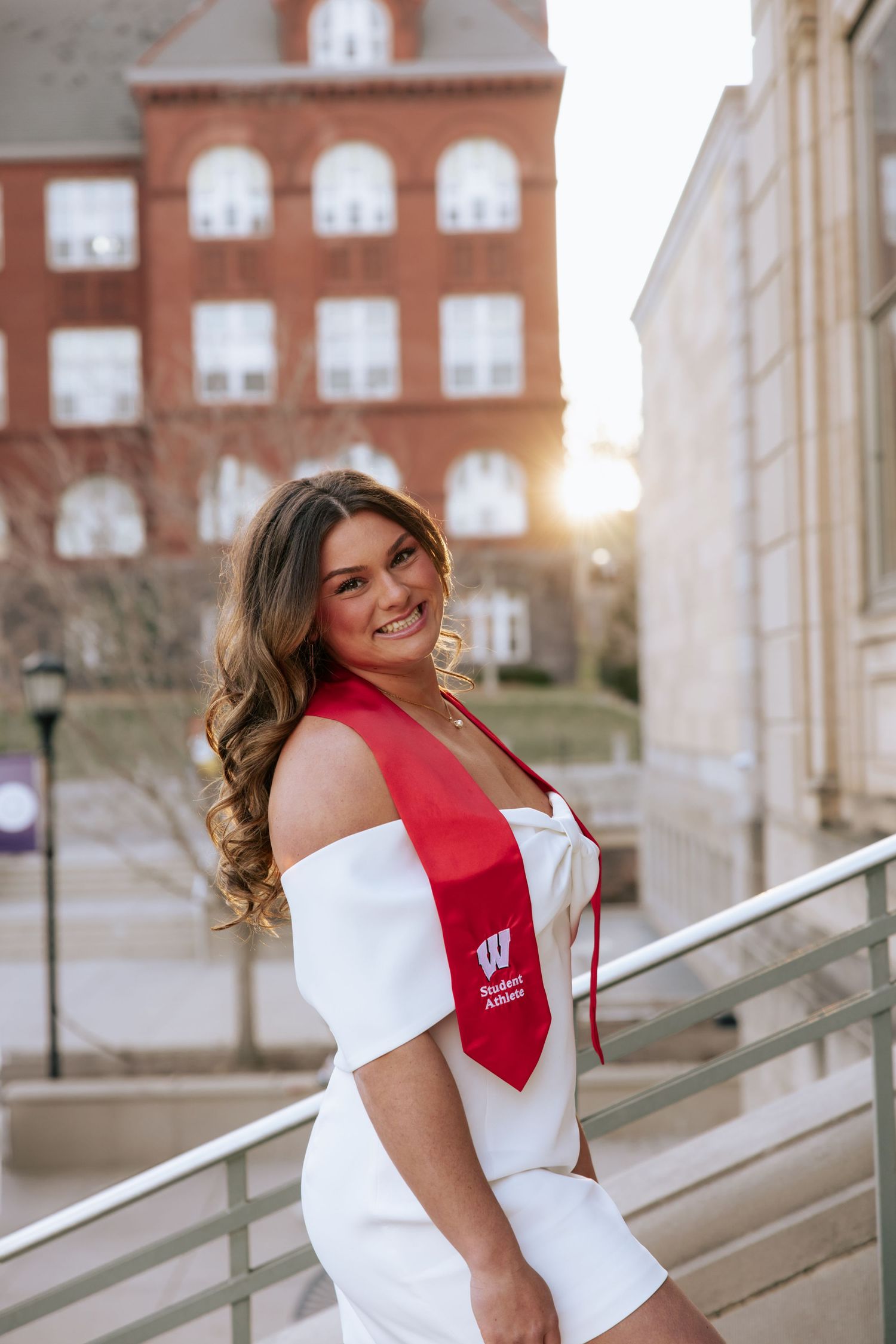Graduate in red and white graduation attire poses on campus steps at sunset with Bascom Hall in background.