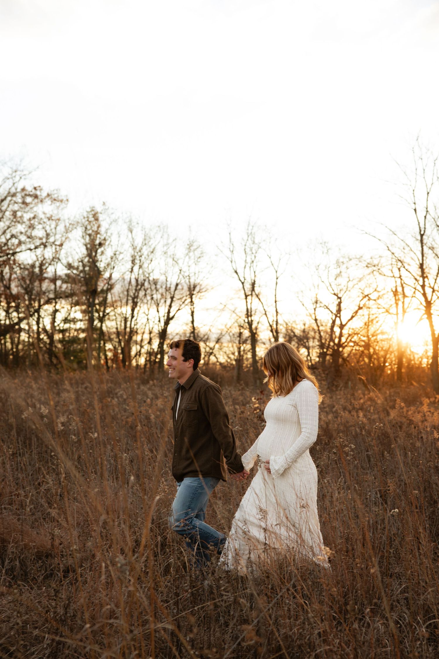 A couple walks through a golden field at sunset holding hands.