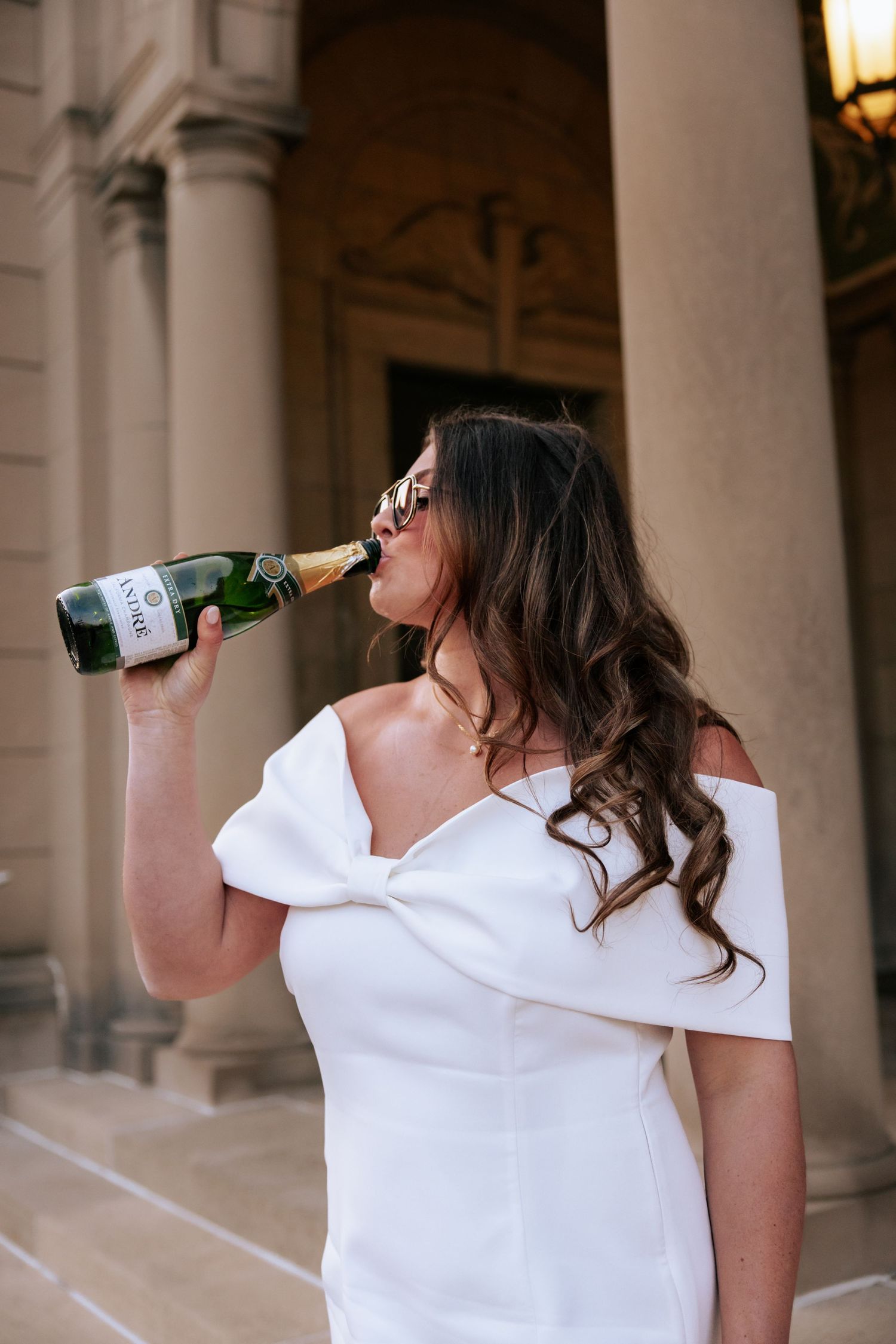 Graduate in white dress celebrating with champagne in front of classical columned building.