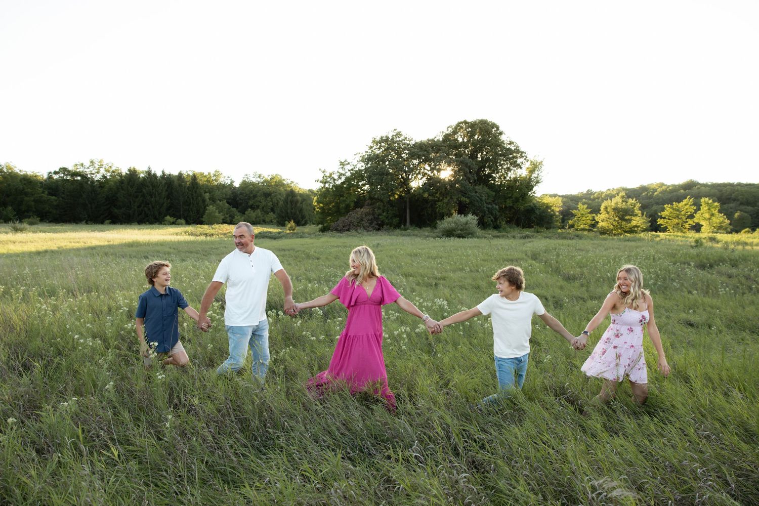 Family holding hands while walking through tall grass in a summer meadow at sunset.