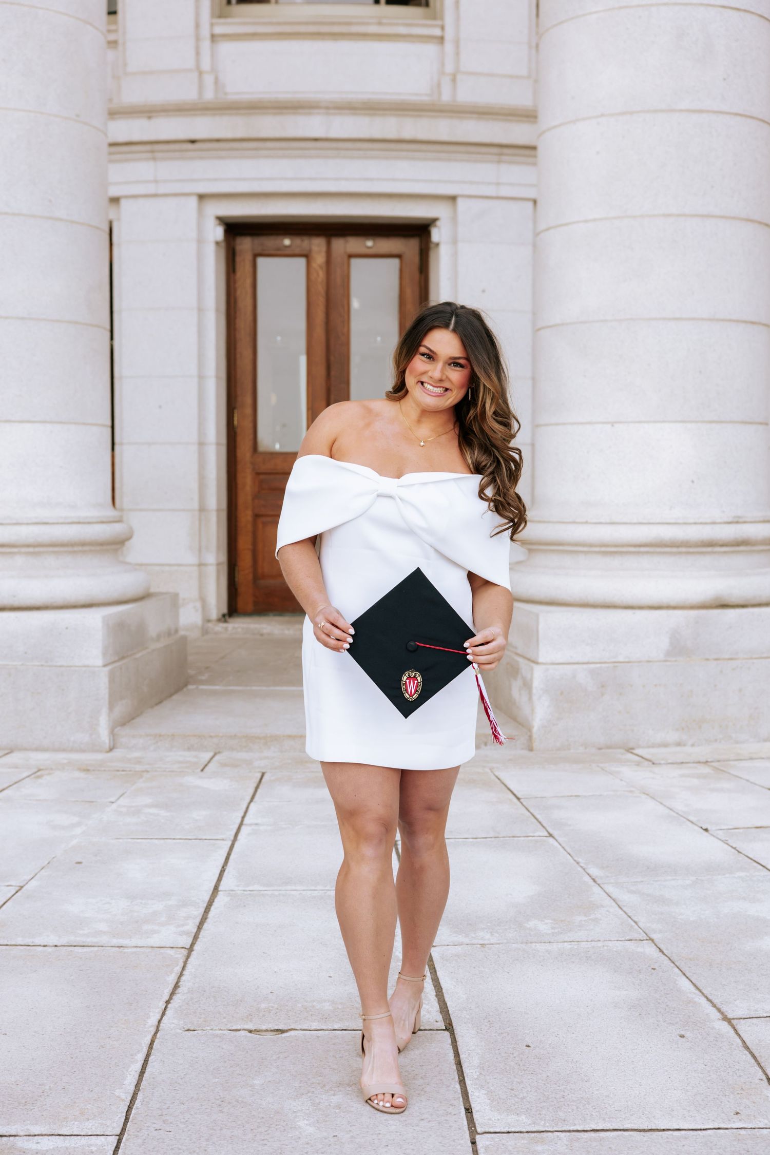 A graduate in a white off-shoulder dress poses with their diploma in front of white stone columns.